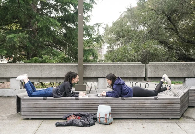 Two people facing each other work on their laptops on a bench outside of Moffitt Library.