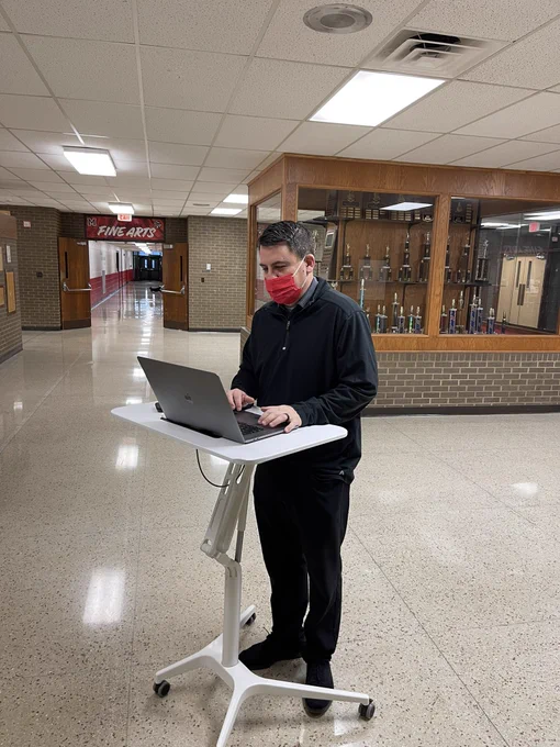 A principal standing at a rolling desk in a hallway, working on a laptop.