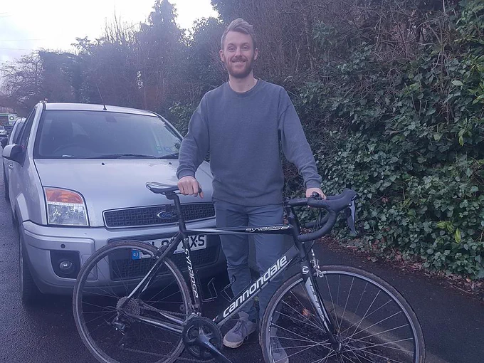 Cyclist with bike next to cars parked on road and petrol garage in background