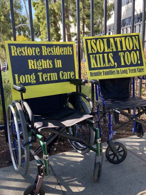 2 wheelchairs sit beside each other in front of metal bars. The wheelchairs are empty other than large yellow and black signs. The sign on the left reads: Restore Residents in Long Term Care. The sign on the right reads: Isolation Kills, Too!