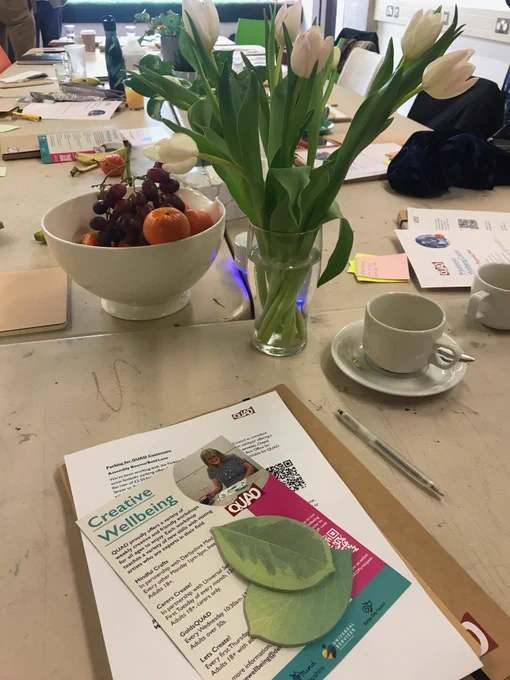 A table with a coffee cup, fruit bowl, leaf shaped post it notes, a pen and creative wellbeing leaflets on. At the centre of the table is a vase of white tulips.