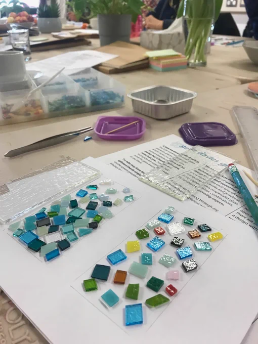 A table with a sheet of white paper on. On the paper are two glass oblongs with glass mosaic patterns on, that have been created.