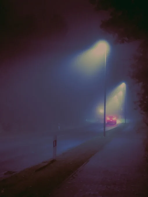The German city Geldern in the lower rhine area at night. A street winds from the left to the distance. It is foggy and there are only a few street light sources visible. The street lights are slightly blue and in the center right of the image on the street there is a car hitting the breaks, showing its red rear lights. In the foreground left there is a road delineator visible in the dark. Some dark foliage is visible on the right. The image looks outerworldy and dreamy.

Photo by Constantin Flux Photography. constantinflux.com