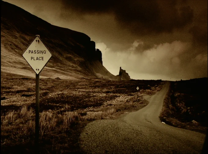 Film Still from The Call of Mist (Redux) featuring an empty road through a mountain pass with a sign that reads Passing Place