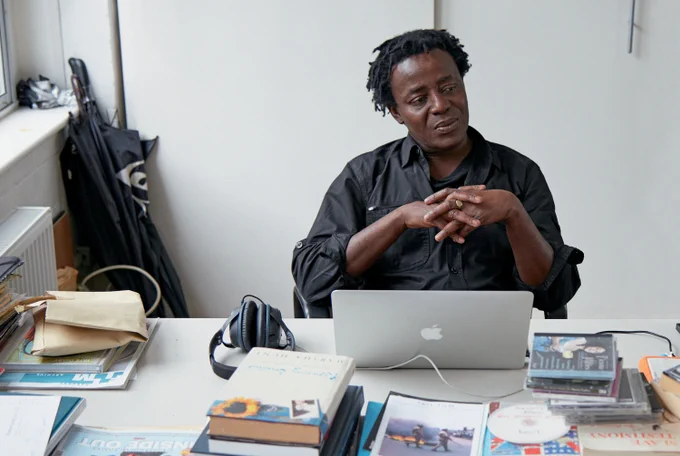 John Akomfrah sits by desk with laptop with hands folded at chest