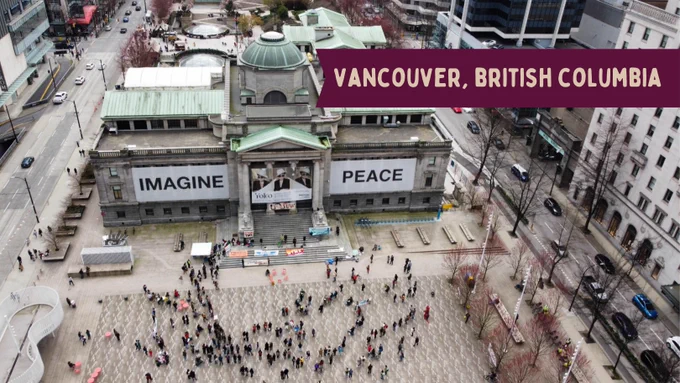 A group of activists gather around a building with a banner hung on it that reads "imagine peace". Yellow text on a red label at the top right reads "Vancouver, British Columbia"