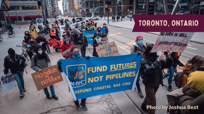 A group of activists march on the street behind a banner with the Wet'suwet'en bear attacking the RBC lion logo and text that reads "fund futures not pipelines defund coastal gas link". They hold various signs that read "no nature no future" and "uproot the system". Photo is by Joshua Best. Yellow text on a red label at the top right reads "Toronto, Ontario"