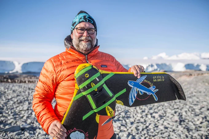 Dr Dave Johnston in Antarctica, penguins and icebergs in the background, stands facing the camera holding a drone.
