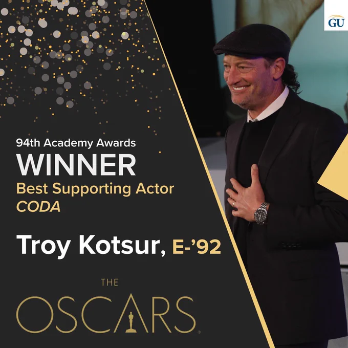 [Image Description: dark grey asymmetrical banner with buff outline. Gold colored confetti falls from the top. Text reads: “94th Academy Awards Winner, Best Supporting Actor, CODA. Troy Kotsur, E-‘92. The Oscars logo. An image of Troy, a white man wearing a black hat and black suit rests his hand on his chest, smiling.]