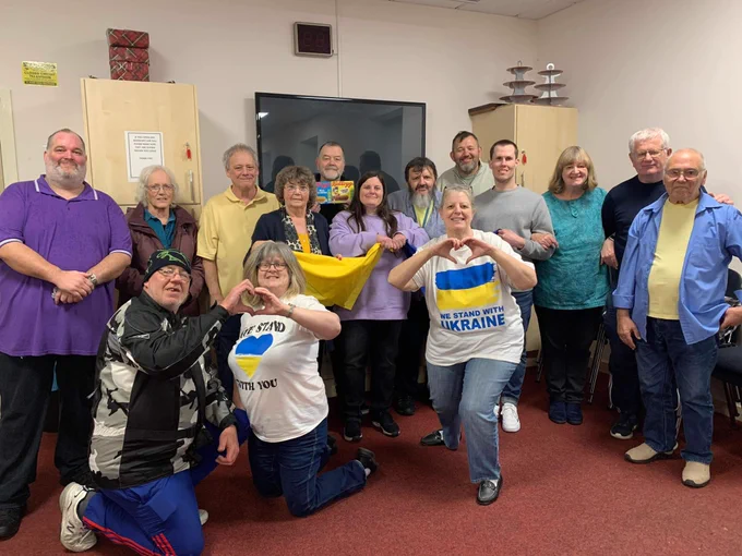 A group of men and women smiling for the camera whilst wearing ‘we stand with Ukraine’ T-shirts. Some have used hand shapes to create a love heart gesture.