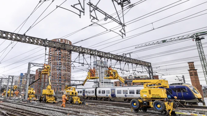Overhead electric wires being upgraded above the railway at Leeds Station while a Northern train passes behind. Candle House, a tall cylindrical building, is in the background.