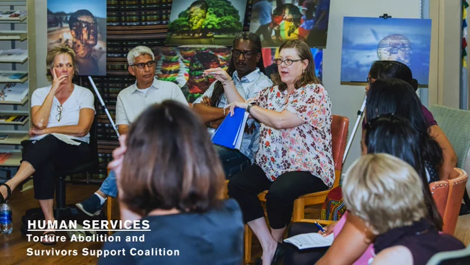 Photo of a woman wearing blue glasses and a floral print top speaking with a circle of other people. In the background, pieces of art are attached to large whiteboards. White text at the bottom reads: Human Services, underlined, Torture Abolition and Survivors Support Coalition.