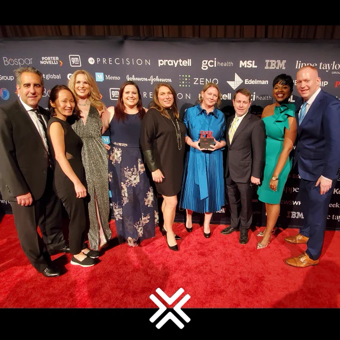Group photo of 9 current global staff members all in formal attire, standing on the red carpet at the PRWeek awards show. Behind them is a banner with sponsors of the show. 