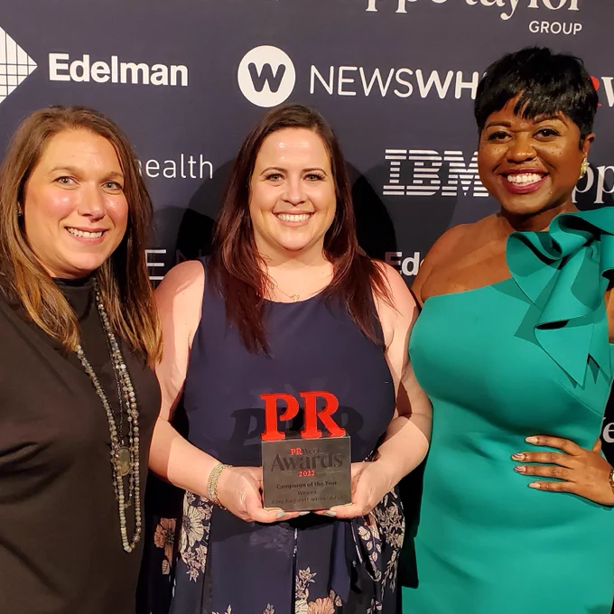A photo of three CG staff members: Mara Mazzoni, a white woman with brown hair in a black dress; Shannon McGovern, a white woman with brown hair in a blue dress, holding a PRWeek  trophy; Rebecca Roussell, a Black women with short black hair and in a teal dress. Behind them is a banner with sponsors for the awards. 