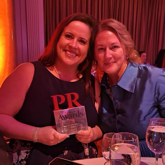 A photo of Shannon McGovern, a white women with medium length brown hair in a black dress holding a PRWeek trophy, and Virginia Devlin, a white women with medium length blonde hair in a blue blouse, sitting at a table with a curtain and lights behind them and some water glasses on the table. 