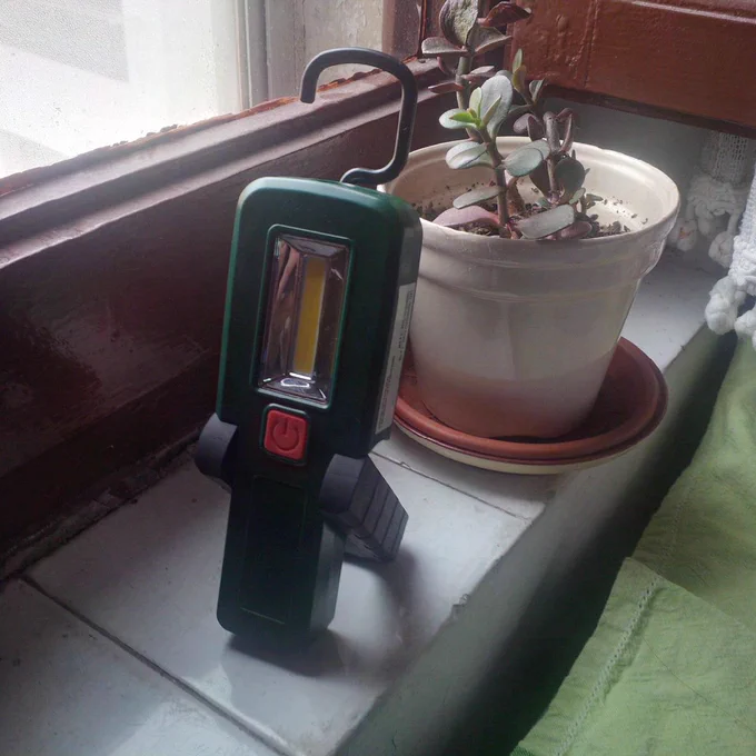 Linterna de pie en una superficie plana, gracias a un pie de goma que se despliega por detrás como soporte. También está desplegado un ganchito hacia arriba para colgarla de cualquier lugar. Se aprecia que es pequeña, no más alta que un tiesto pequeño que hay detrás.