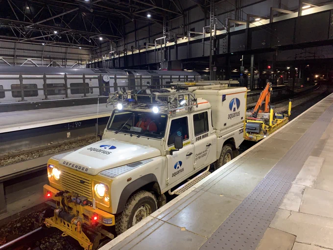 A Land Rover vehicle, fitted with road wheels and rail wheels, runs along the railway track through platform 8 at Leeds Station during the night. The station is empty, apart from an out of service train parked overnight in the background.