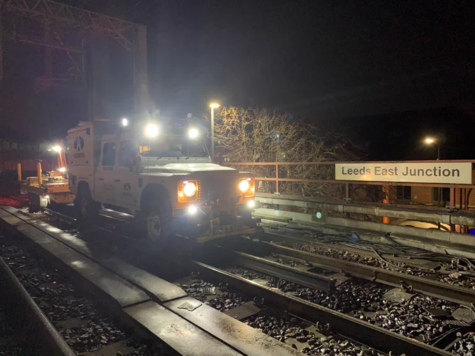 A photograph of the road and rail Land Rover on the railway track in front of a large sign reading Leeds East Junction. It is night time and the Land Rover has its headlights and floodlights switched on.