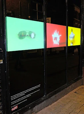 A picture of a shop front, with three windows. The windows have been blacked out. In the centre of each window is a brightly coloured background rectangle with a single image pictured. The first window has a green picture with an image of sunglasses. The second window has a red background with an image of a shell and the final window has a yellow background, with an image of a ceramic pot. 