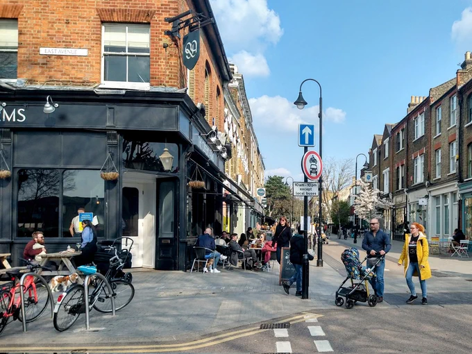 Lots of people walking, sitting and milling about on Orford Road in Walthamstow including sitting outside a pub and cafés. A sign says no motor vehicles in the foreground.