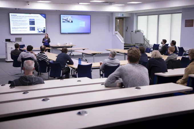 View from the back of the presentation room with Alys at the front and two screens showing slides. You can see the backs of about 13 audience members strewn across the room.