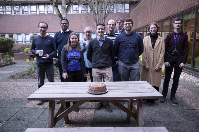 Some RSE Culham team members and friends stand behind a chocolate cake on a picnic table.