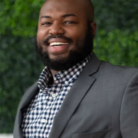 Photo of Brandon Ritter, African-American man with beard, shaved head. Smiling, green background, gray jacket, checked shirt.