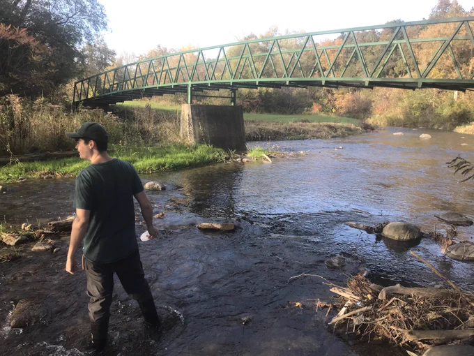 master's student Simon Pollard stands in the Oshawa Creek with a bottle in his hand. There is a green footbridge that spans across the creek, which is lined with trees. 