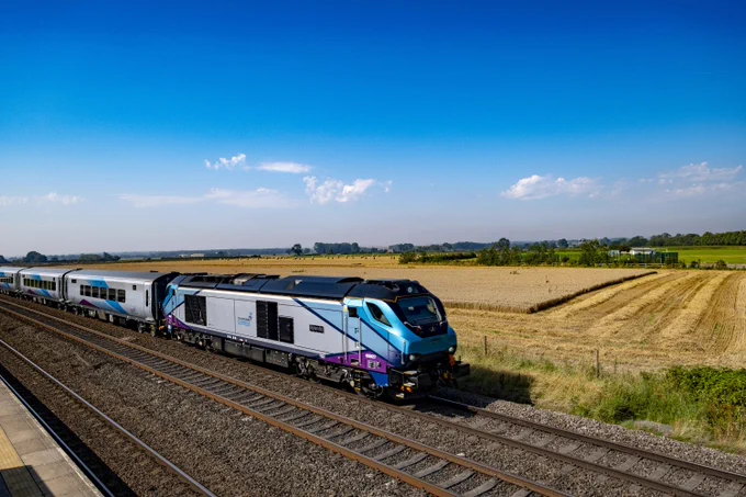 A silver, blue and purple Transpennine Express train speeds past a golden cornfield in the countryside. The weather is sunny and the sky is blue.