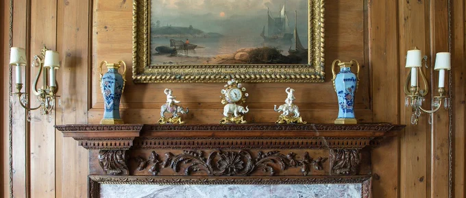 A close-up on a carved, dark wood mantle with a marble fireplace surround and objects on the mantle.