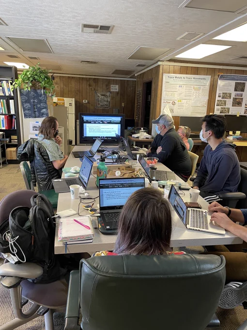 5 members of the OG Fish Lab sit around a long rectangular table with their laptops open. They are all watching a screen playing virtual conference talks from the 2022 IEP Workshop.