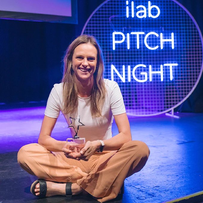 Image UQ ilab Entrepreneur of the Year award recipient, Siobhan Coster sitting on a stage with a trophy in her hands. There is a neon sign in the background which reads: 'ilab PITCH NIGHT'. 