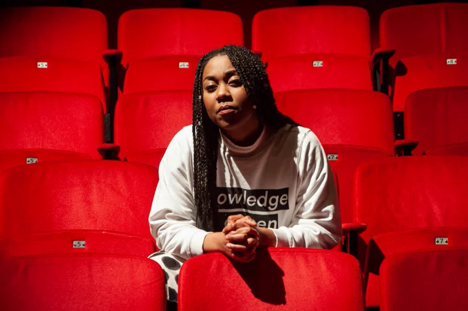 Keisha Thompson sits in a grey sweatshirt in the bright light of a theatre, perched on red chairs and leaning over with her hands clasped and looking down the camera. She has a strong, determined expression on her face.