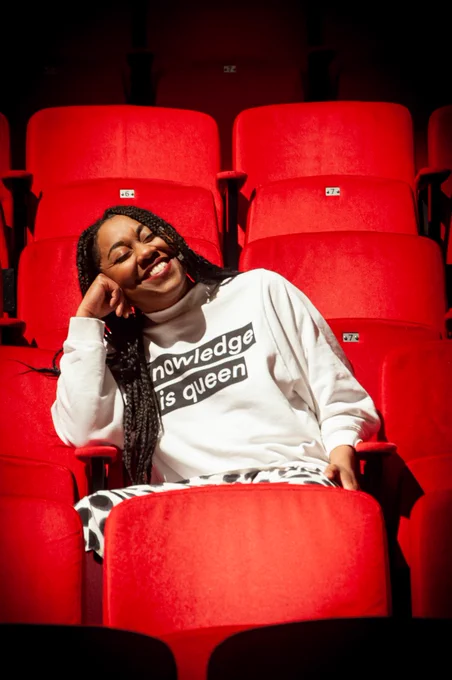 Keisha Thompson leans back in a grey sweatshirt in the bright light of a theatre, perched on red chairs and resting her head on her fist, eyes closed and a joyful smile on her face.