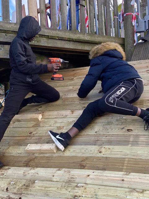Two people leaning against a wooden structure. One has a drill in his hand