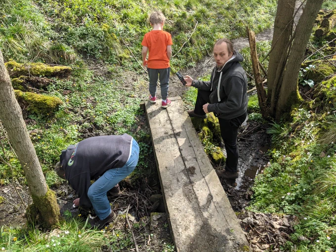 Paul, John and Dan looking around for a geocache container, standing around a bridge that goes over a small stream. Dan and Paul are stood in the stream