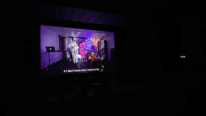 A cinema screen in a dark room. On the screen is Spirit Sigh/Jessica Ashman's Dawta – a room with two musicians performing, one holding a guitar, one sitting at a drum kit. Laid over them is a projection of an animation showing a young black girl. The image of the girl is stylised, cut up, disjointed. Captioned lyrics along the bottom of the screen read: I don't know who I want to be.