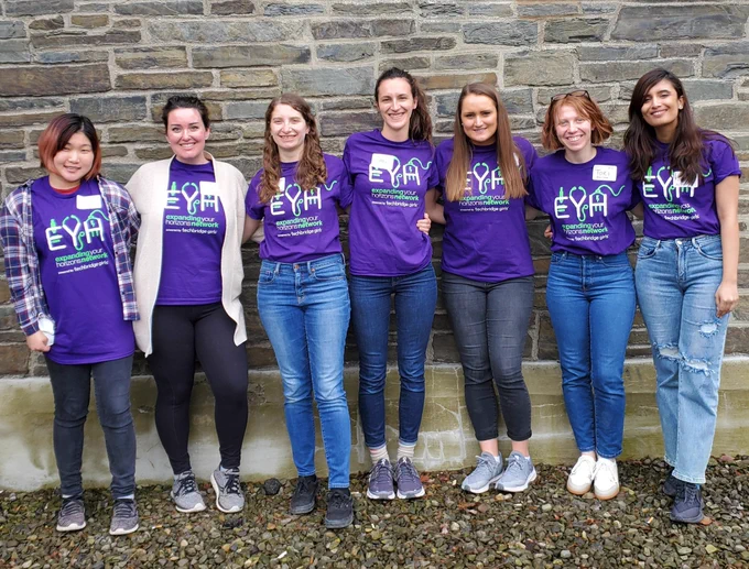 7 adult females are standing up and smiling towards the camera. They are all wearing purple t-shirts with "expanding your horizons network EYH". In the background, there are bricks of the building.