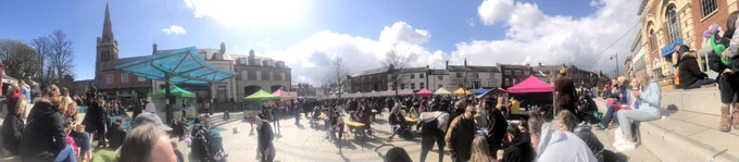 Kettering Market place full of people eating, sitting, walking and watching musician, lots of food stalls ,sunshine, church spire and old buildings in background.