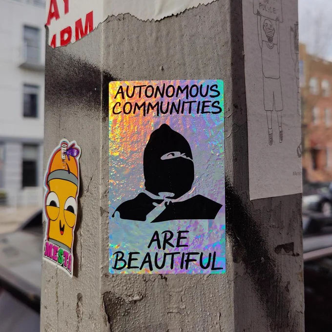 Shiny sticker on a pole featuring a Zapatista and the words Autonomous Communities Are Beautiful"