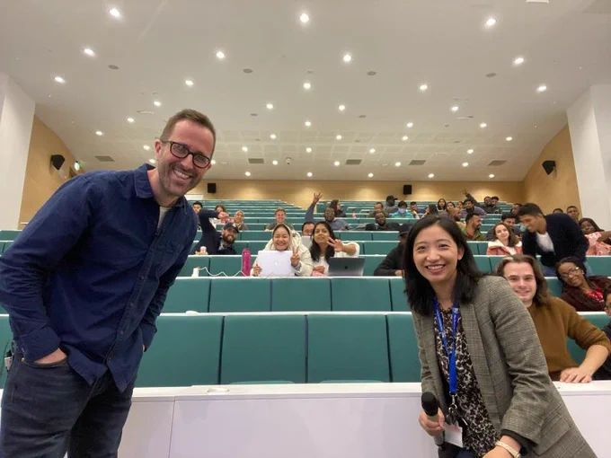 Tom Quay and Dr Danni Liang smiling at the camera in front of a lecture theatre full of students, who are smiling and waving. 