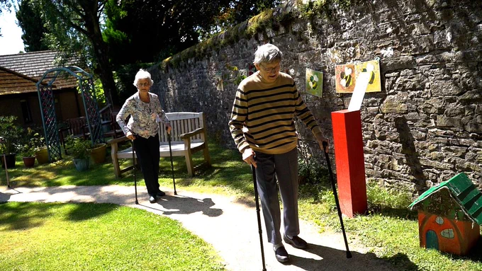 Two ladies walking in a care home garden on a path using two walking sticks each.