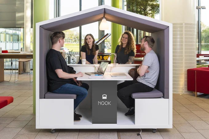 Photo of a white Nook pod with grey interiors. There are two people sat inside, talking to two people on the other side, as the back panel has been removed. 