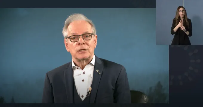 An aging man wearing eyeglasses is sitting on a sofa chair in front of a step-and-repeat banner with the text Canada Council for the Arts. Bringing the Arts to Life. He is speaking into a camera.