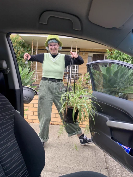 Photo of Is, a white trans person. They wear green pants, a green vest, black turtleneck and a green hat. They have blue short hair, glasses and are grinning happily. They are holding 2 plants in their arms and are outside an open car door.