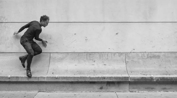 A black and white image of the artist, a Black man, dressed in dark clothing, climbing on a sloped ridge along an exterior wall. One hand rests against the wall, and one foot is elevated as if he is in motion
