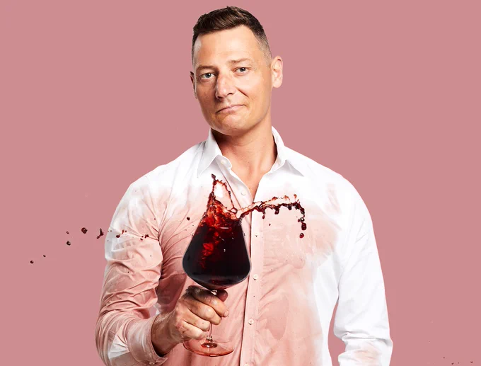 Merrick Watts holds up a glass of wine that spilled on his white shirt. He has brown hair and stands in front of a pink background.