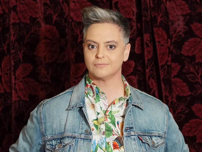 Geraldine Hickey smiles at the camera. She wears a Hawaiian shirt with a denim jacket over the top. She has grey hair and stands in front of a red velvet curtain.