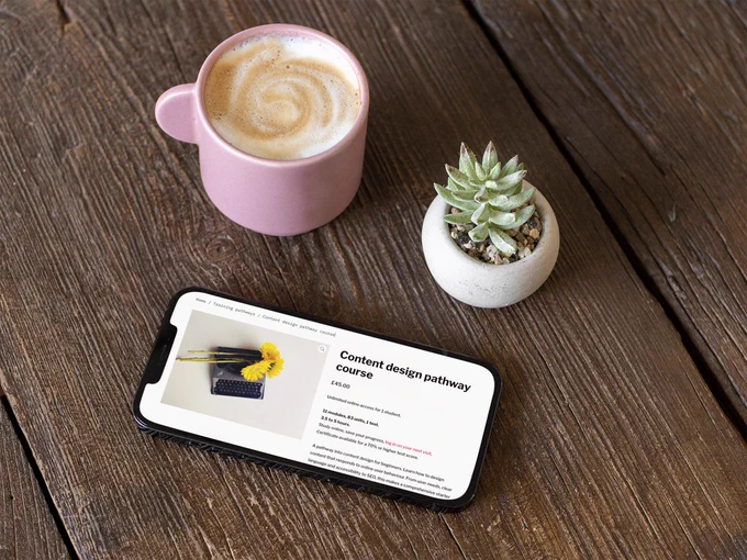 Pink mug of latte, succulent and mobile phone on a wooden plank tabletop. The mobile screen shows Content design pathway product page with a decorative image of a vintage typewriter with sunflowers laid on it.