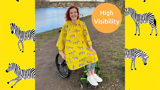 Alt: A picture of a person in a wheelchair with red hair. They are wearing a bright yellow poncho with zebras on it. In the background is a river.